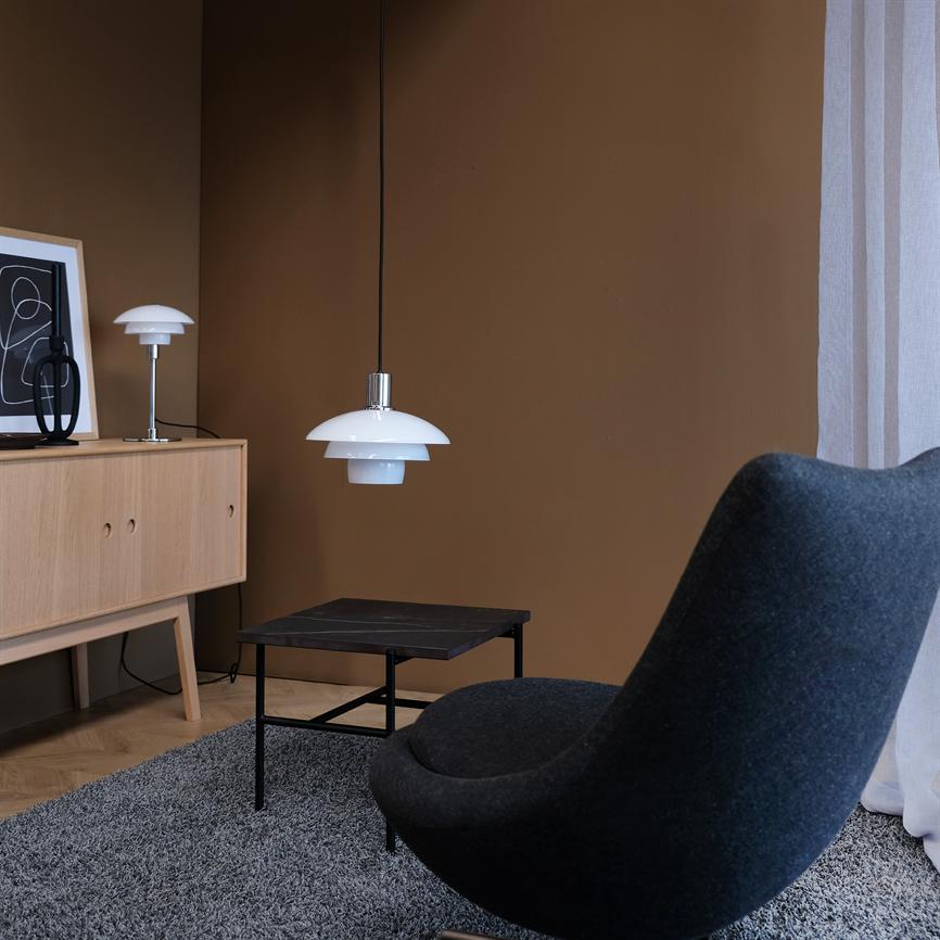 Weiß pendel hängt über einem niedrigen schwarzen Couchtisch in einer minimalistischen Wohnzimmerecke mit einer braunen Wand, einem Schrank aus heller Eiche, einer Tischlampe und einem dunkelgrauen, geschwungenen Sessel.