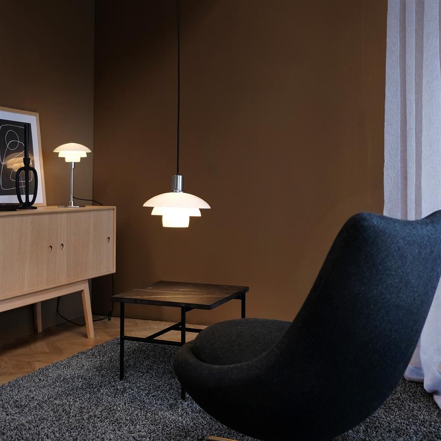 Hängelampe leuchtet sanft über einem kleinen Couchtisch; umgeben von einem schwarzen, geschwungenen Sessel, grauem Teppich, hellem Holzschrank und dunkler Wand von einem hellen Vorhang.