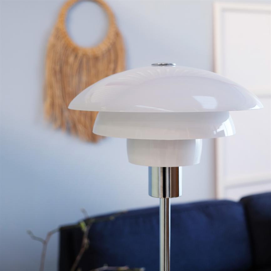 Eine weiße, glänzende Tischlampe steht unbeleuchtet auf einem Chromstab vor einem dunkelblauen Sofa in einem hellen Raum mit einer Wanddekoration aus Korbgeflecht und einem schmalen Bilderrahmen an der Wand.