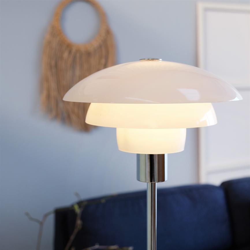 Eine moderne weiße Stehlampe leuchtet sanft; Es steht auf einem Chromständer vor einem dunkelblauen Sofa, dahinter befindet sich eine hellblaue Wand, die mit Wanddekoration aus Korbgeflecht und einem Bilderrahmen verziert ist.