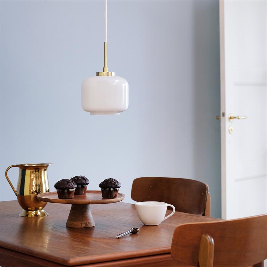 Über einem Holztisch hängt eine weiße Glaspendelleuchte; Auf dem Tisch steht ein Kuchenteller mit drei Schokoladenmuffins, ein goldener Krug und eine Tasse in einem einfachen Esszimmer mit hellblauen Wänden und offener Tür.