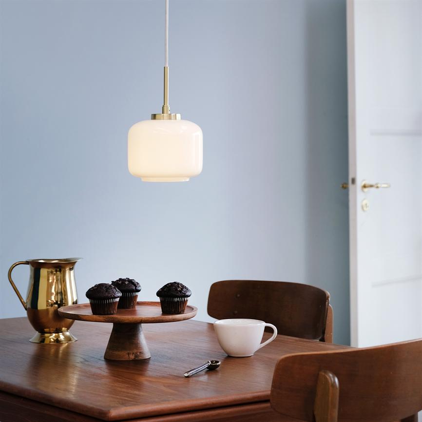 Über einem Holztisch hängt eine weiße Glaspendelleuchte mit drei Schokoladenmuffins auf einem hölzernen Kuchenständer, einer weißen Tasse, einem goldenen Krug und Holzstühlen vor einer hellblauen Wand und einer offenen Tür.