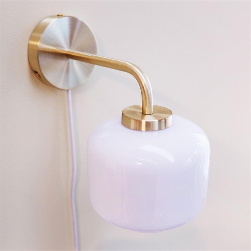 Wandleuchte mit opalweißer Glaskuppel hängt an einem gebogenen Messingarm, montiert auf einer runden Wandplatte; Die Lichtquelle zeigt nach unten in Richtung einer hellen Wand, mit einem sichtbaren weißen Draht.