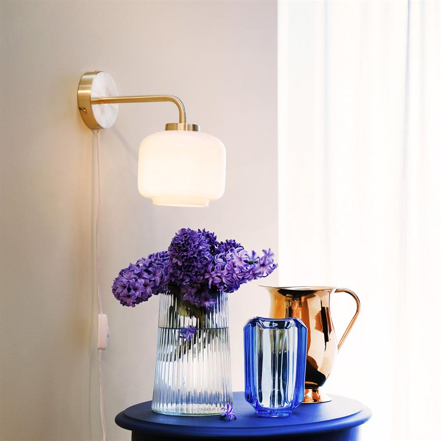 Der Strauß violetter Hyazinthen steht in einer klaren geriffelten Glasvase, beleuchtet von einer Wandleuchte, flankiert von einer blauen Glasvase und einem Kupferkrug auf einem blauen runden Tisch vor einem Lichtvorhang.