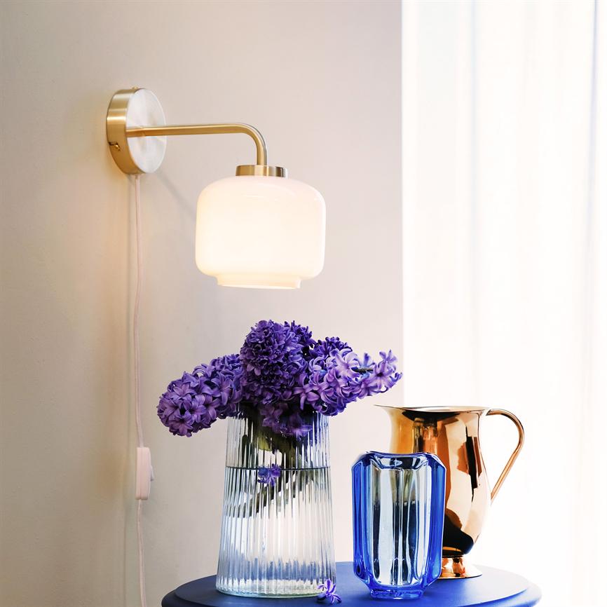 Ein Strauß violetter Hyazinthen steht in einer geriffelten Glasvase auf einem runden bläulichen Tisch; Eine weiße Wandlampe beleuchtet sie vor einem hellen Vorhang, flankiert von einer blauen Glasvase und einem Kupferkrug.