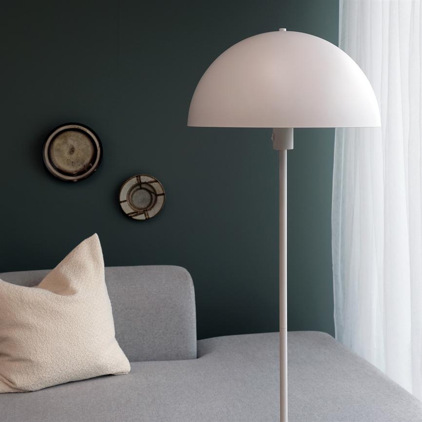 Neben einem grauen Sofa steht eine weiße Stehlampe; Auf dem Sofa liegt ein cremefarbenes Kissen, im Hintergrund eine dunkelgrüne Wand mit zwei Dekotellern und weißen Vorhängen.