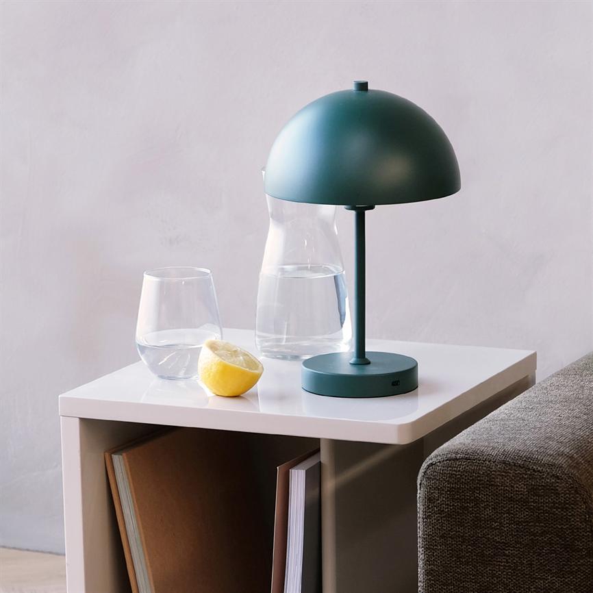 Auf einem weißen Beistelltisch steht eine grüne Tischlampe; daneben liegen eine Glaskaraffe, ein Glas Wasser und eine halbe Zitrone; Der Tisch steht neben einem grauen Sofa vor einer hellen Wand.