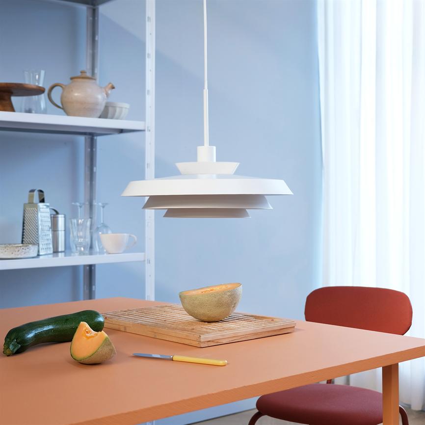 Eine weiße Pendelleuchte hängt über einem terrakottafarbenen Tisch, auf dem in einer hellen Küche eine halbgeschnittene Melone auf einem Schneidebrett, ein Kürbis, ein Messer und ein roter Stuhl stehen.