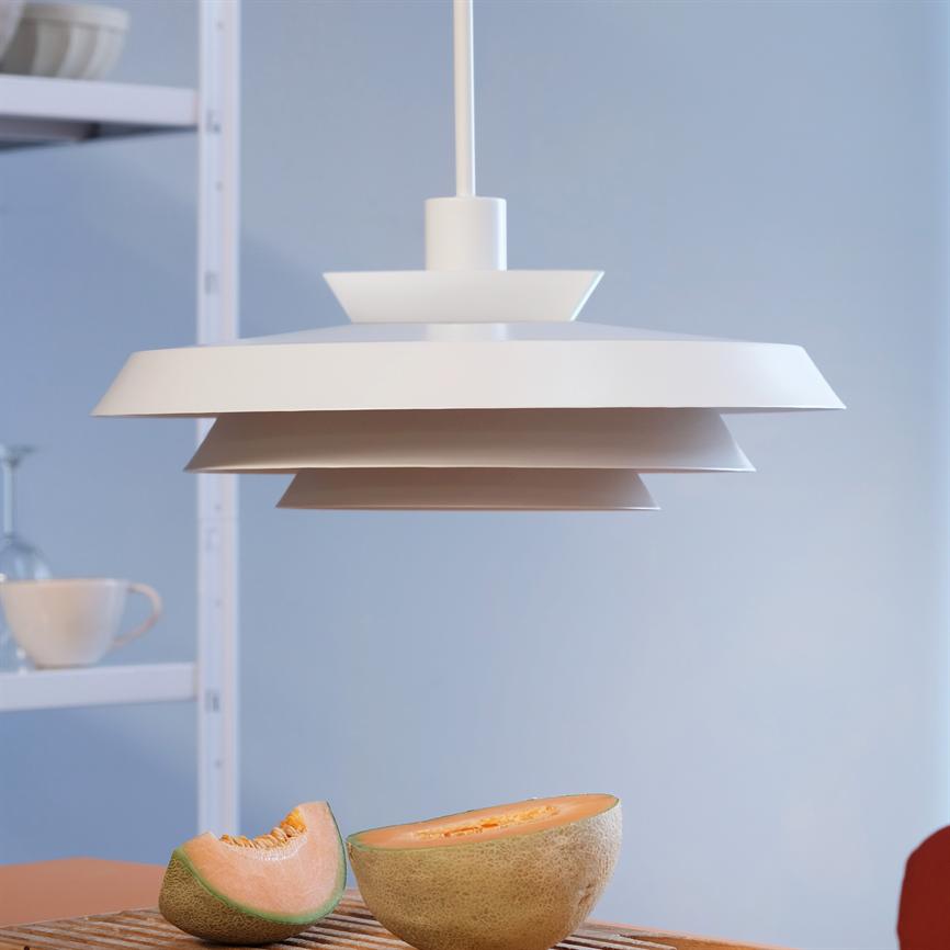 Objekt: Weiße, geschichtete Pendelleuchte. Aktion: Leise von der Decke hängen. Kontext: Heller, minimalistischer Küchen-/Esstisch mit einer halbierten Cantaloupe-Melone auf dem Tisch und einem Regal mit einer Tasse im Hintergrund.