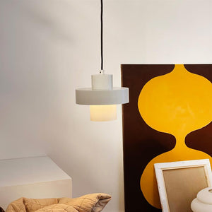 Eine weiße Hängelampe hängt an einer schwarzen Schnur und strahlt sanftes, warmes Licht aus; es schwebt vor einem großen Gemälde mit gelber abstrakter Form, umgeben von Rahmenstapeln und einem beigen Teppich.