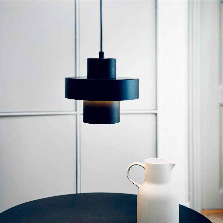 Schwarz pendel- Die Lampe hängt und wirft sanftes Licht über einen runden, dunklen Tisch, auf dem ein weißer Keramikkrug steht. minimalistischer, heller Raum mit geteilten Wandpaneelen im Hintergrund.