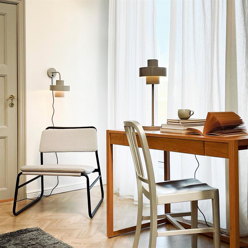 Skrivebordet bærer en åben bog, stak bøger og en kop, mens en bordlampe er tændt; en metalstol og en polstret stol står i et lyst rum med hvide gardiner.
