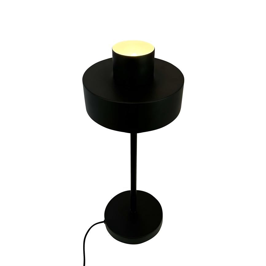 Schwarze Tischlampe mit zylindrischem Schirm, die ein warmes Licht nach oben abgibt; dünner Stab verbindet Bildschirm mit rundem Fuß, sichtbarer schwarzer Draht, vor einem einheitlichen weißen Hintergrund.