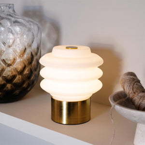 Tischleuchte mit geripptem Opalglas und Messingsockel spendet sanftes, warmes Licht auf einem Regal neben einer gemusterten Glasvase und einer Schale mit Schnur.