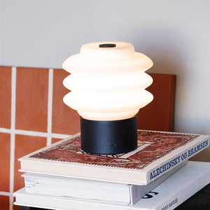 Lampe auf Bücherregal: Eine weiße, gewellte Tischlampe leuchtet sanft und steht auf zwei Kunstbüchern an einem Tisch mit Terrakottafliesen im Hintergrund. Text auf dem Rücken: „ALECHINSKY AND BRAMSEN“.