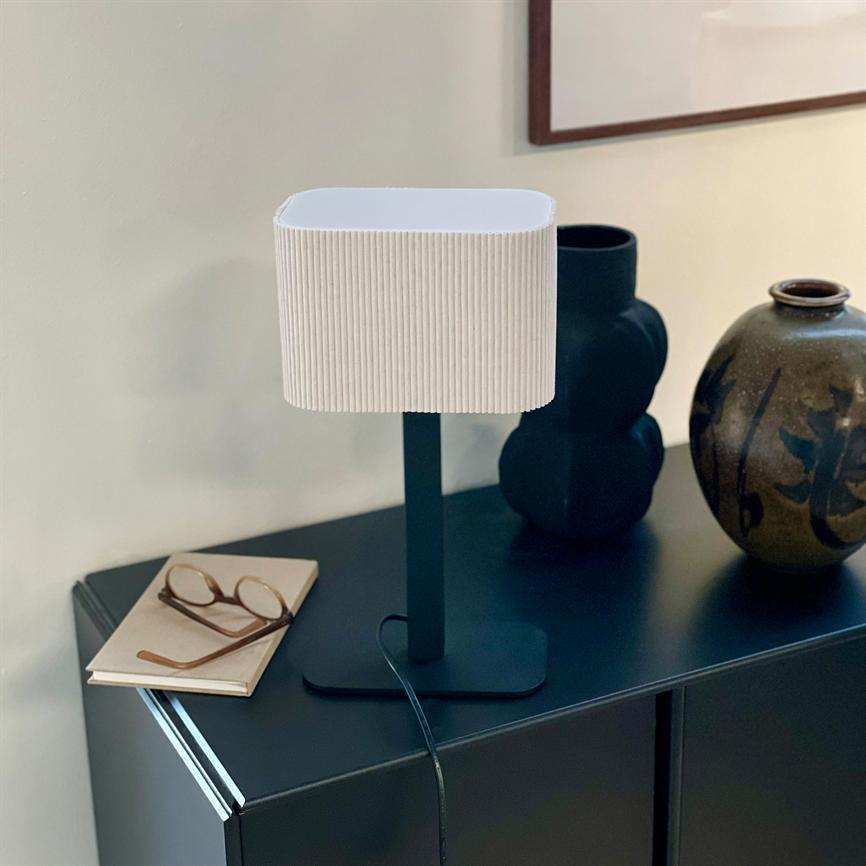 Bordlampe med hvid, rillede lampskærm står på en mørk skænk; ledning løber ned, et par briller ligger på en notesbog til venstre, to keramiske vaser står bagved.