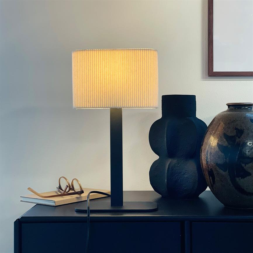 Tischleuchte strahlt warmes, goldenes Licht aus; Es steht auf einem dunklen Schrank zusammen mit einer schwarzen Vase, einem glasierten Glas und einer Brille auf einem Notizbuch unter einem gerahmten Poster.