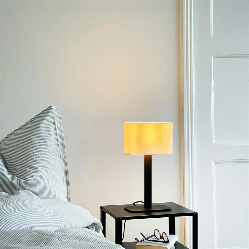 Auf einem schwarzen Nachttisch brennt eine Tischlampe und wirft warmes gelbes Licht; daneben eine Brille und ein Buch, daneben ein gestreiftes Kissen und eine weiße Tür in einem hellen Schlafzimmer.