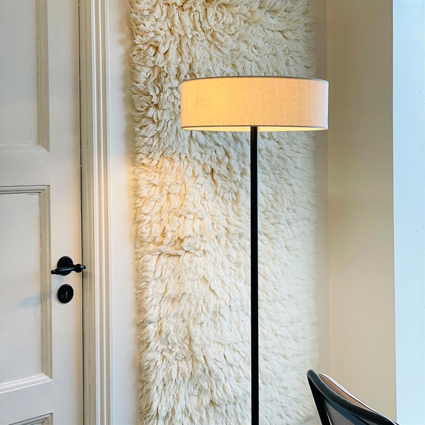 Gulvlampe står tændt i et hjørne foran et stort cremefarvet, uldent vægteppe; til venstre en hvid dør med sort dørhåndtag, og nederst til højre ses en stoleryg.