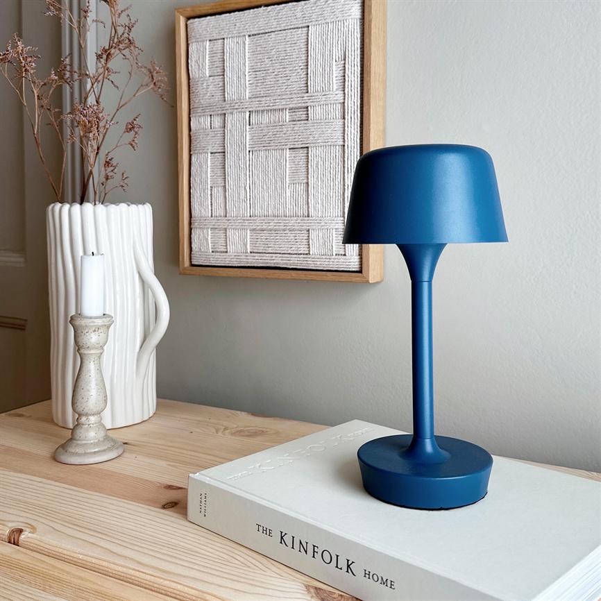 Eine blaue Tischlampe steht auf einem Buch, umgeben von einer hellen Holzkommode mit weißer Vase, Kerzenhalter und gerahmter gewebter Kunst an einer neutralen Wand. Text: „THE KINFOLK HOME“.