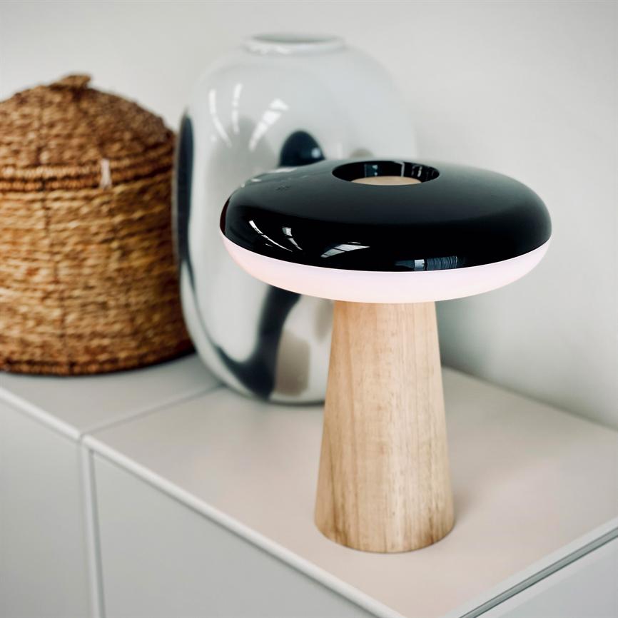 Tischleuchte mit Holzsockel und rundem, schwarz glänzendem Schirm strahlt sanftes rosa Licht aus; steht auf einer weißen Kommode vor einer glänzenden silbernen Vase und einem Weidenkorb in einem hellen, minimalistischen Raum.