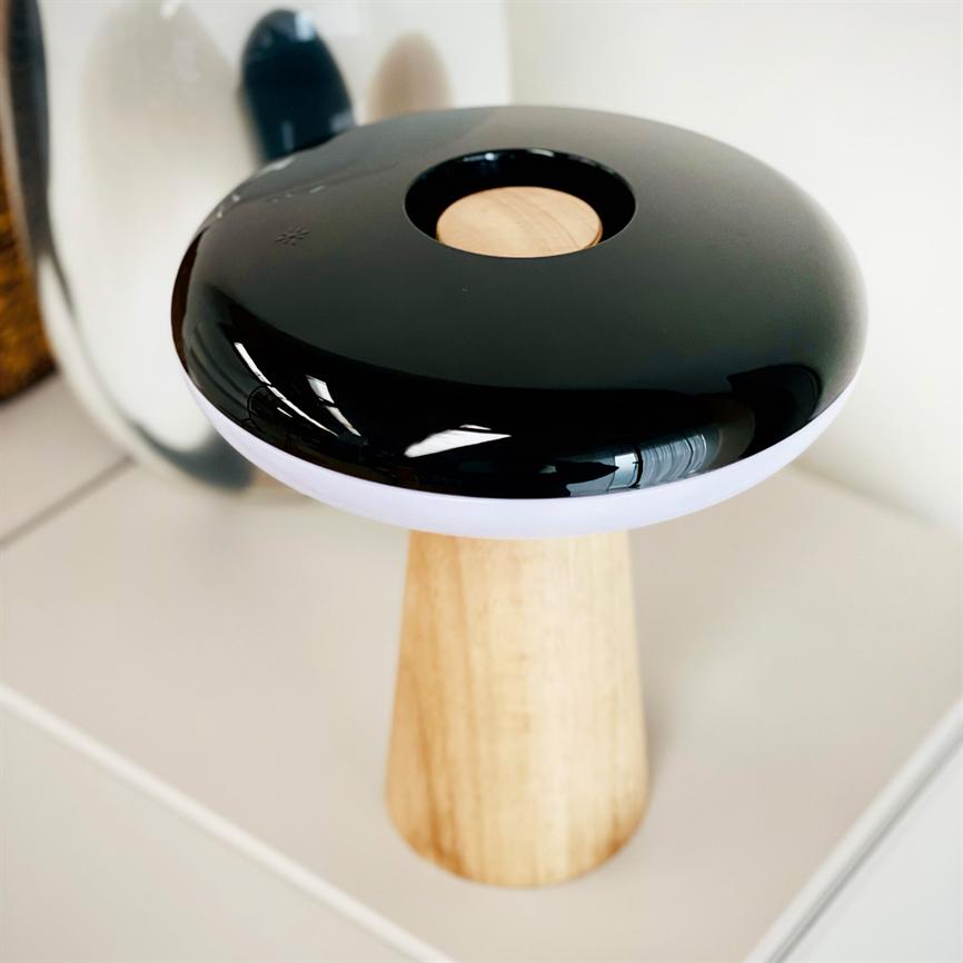Eine schwarze, glänzende, pilzartige Lampe mit rundem Holzeinsatz und weißem Rand steht auf einem hellen Holzsockel auf einer weißen Tischplatte und spiegelt sich in einem Spiegel dahinter.