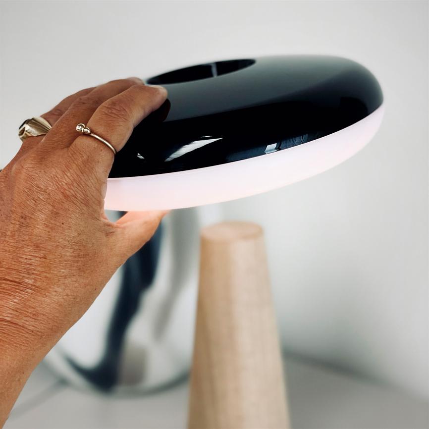 Tischleuchte – eine Hand hebt den schwarzen, runden Schirm mit leuchtend weißem Rand an; Die Lampe steht auf einem kegelförmigen Holzsockel in einem hellen, minimalistischen Raum.