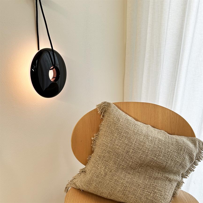 Schwarze Wandleuchte hängt an einer schwarzen Schnur und strahlt warmes Hintergrundlicht aus; Heller Holzstuhl mit rauem beigem Kissen steht vor weißen Wänden und einem weißen Vorhang in einer ruhigen Ecke.