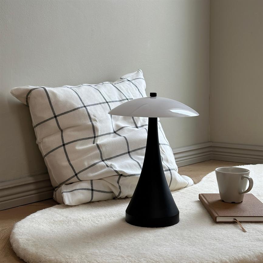 Inmitten eines weichen runden weißen Teppichs steht eine schwarze, kegelförmige Tischlampe mit weißer Kuppel; An der Wand lehnt ein kariertes Kissen, daneben ein Notizbuch und eine Kaffeetasse.