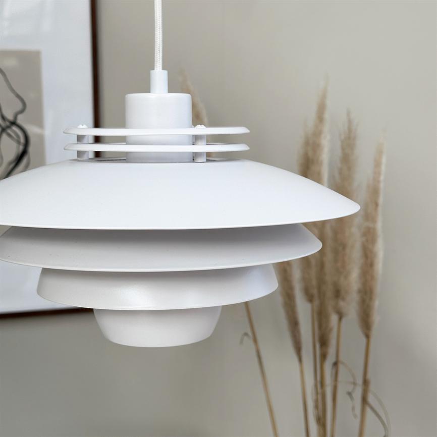 Ein Weiß, mehrschichtig pendel Lampe hängt an textilummantelter Kordel; Es schmückt ein ruhiges Interieur vor einer beigen Wand mit getrocknetem Pampasgras und einem gerahmten abstrakten Druck.