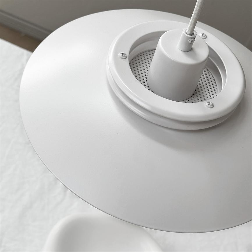 An einer mit Stoff überzogenen Kordel hängt eine weiße Metallpendelleuchte; Der runde Schirm und die perforierte Fassung der Lampe sind schräg zu sehen. Kontext: Helles, minimalistisches Interieur mit weißem Textil und neutraler Wand.