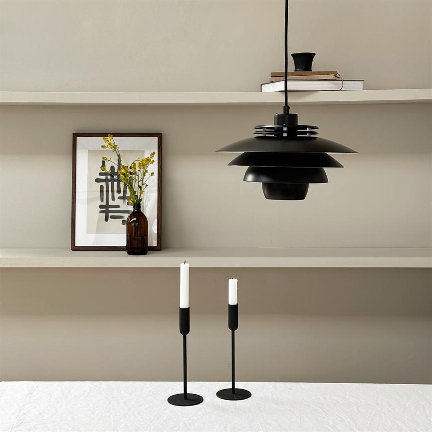 Über einem weiß gedeckten Tisch hängt eine schwarze, abgestufte Pendelleuchte; Auf dem Tisch stehen zwei schlanke schwarze Kerzenständer mit weißen Kerzen. Im Hintergrund Regale mit einem gerahmten Poster und einer braunen Flasche mit gelben Blumen.