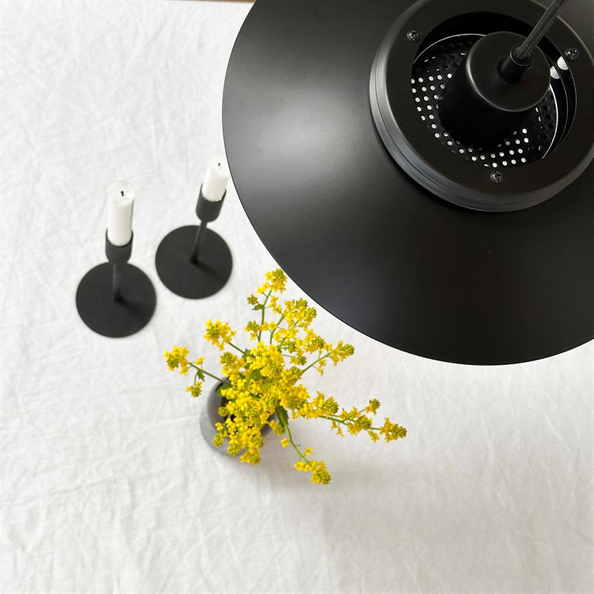 Über einer weißen Tischdecke hängt eine schwarze Deckenlampe, die eine kleine Vase mit gelben Blumen beleuchtet; Auf der Oberfläche der Tischdecke stehen zwei minimalistische schwarze Kerzenständer mit weißen Kerzen.