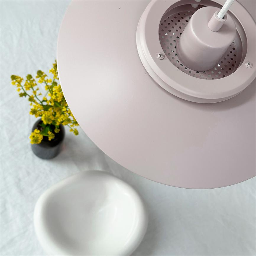 Eine rosafarbene Pendelleuchte dominiert den Vordergrund; Es hängt über einem Tisch mit einer kleinen Vase mit gelben Blumen und einer weißen, ovalen Schale auf einer hellen Tischdecke.