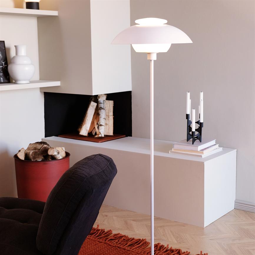 Eine Stehlampe wird von einer niedrigen weißen Bank mit Büchern und einem Kerzenständer beleuchtet; eine kleine Kaminöffnung mit gestapeltem Birkenholz und ein dunkles Sofa und ein roter Teppich im Vordergrund.