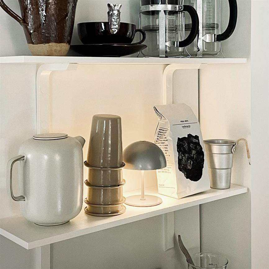 Eine kleine graue Tischlampe leuchtet schwach auf einem weißen Küchenregal, umgeben von einem Wasserkocher, einem Stapel Tassen, einer Kaffeetüte und einer kleinen Metalltasse. 
 Text auf der Tasche: „kathrine“ (teilweise undeutlich) und mehrere kleine, unleserliche Zeilen.