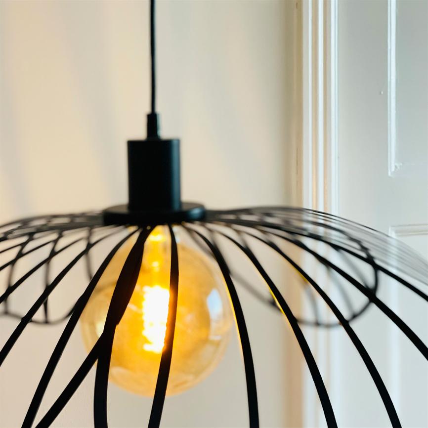 Deckenleuchte mit sichtbarem, leuchtendem Leuchtmittel hängt in einem schwarzen, spinnenartigen Metallkäfig und strahlt warmgelbes Licht aus; umgeben von hellen Wänden und einer weißen Tür im Hintergrund.