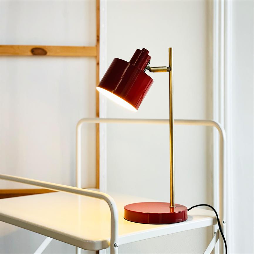 Red desk lamp lights downwards, mounted on a brass rod; es steht auf einem weißen Rollwagenregal aus Metall mit schwarzer Kordel, vor einer hellen Wand und einem schlichten Holzrahmen.