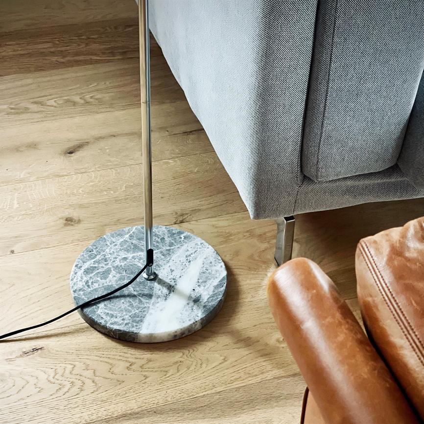 Neben einem grauen Polstersofa steht eine Stehlampe mit rundem Marmorsockel; Chromstab und schwarzer Draht liegen auf einem hellen Holzboden, daneben ein brauner Ledersessel.