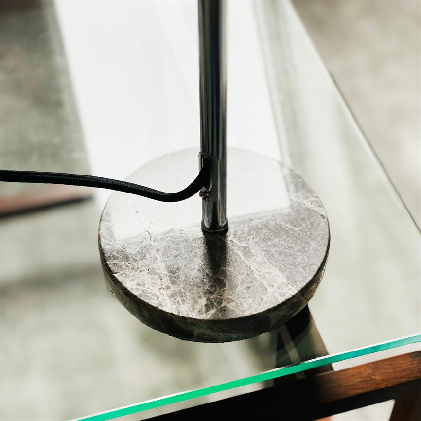 Runder Marmorsockel für Lampe ruht auf einem Glastisch; Aus der Mitte erhebt sich ein Chromstab, aus dem Stab führt eine schwarze Textilschnur heraus; Hintergrund ist verschwommener Boden.