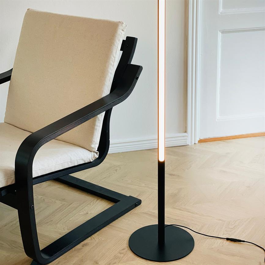 Stol med lyst polstret sæde og sort træramme står ved en slank, tændt gulvlampe på lyst trægulv foran en hvid væg og en dør.