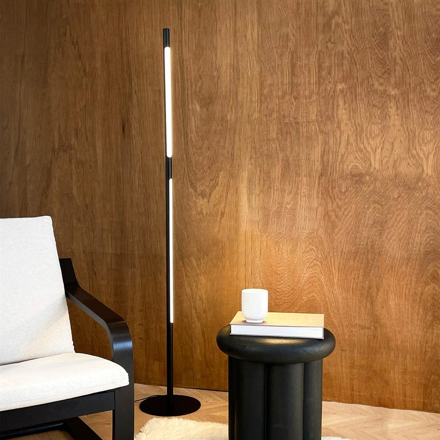 Slank, oplyst gulvlampe står vertikalt og kaster blødt lys over en trævæg; et lyst polstret lænestol til venstre og et sort sidebord med bog og hvid kop til højre.