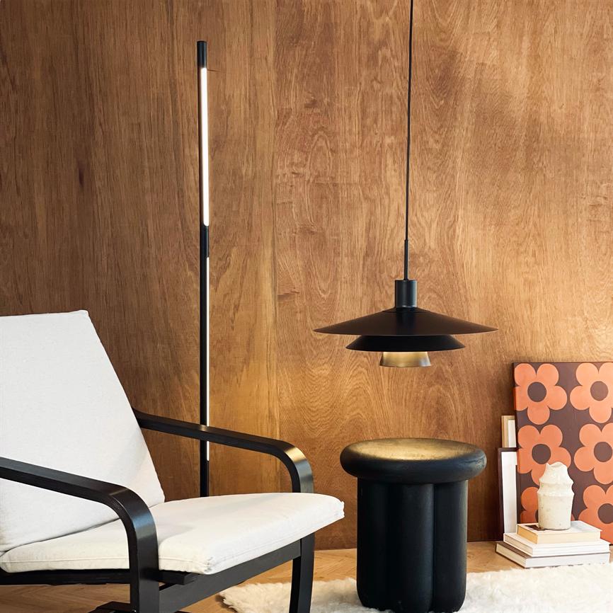 Hvid polstret lænestol står roligt foran en træbeklædt væg; en sort pendel hænger over et rundt sort skammel, flankeret af en slank gulvlampe, plakater, bøger og et lys.