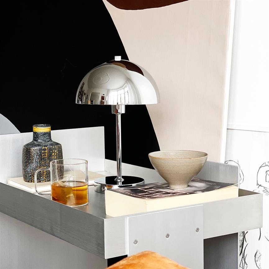 Auf einem silbernen Nachttisch steht eine verchromte Kuppellampe, die den Raum widerspiegelt. drumherum ein Glas Tee, eine gemusterte Vase, eine Schale und ein Buch vor einer abstrakten schwarz-rosa Wand.