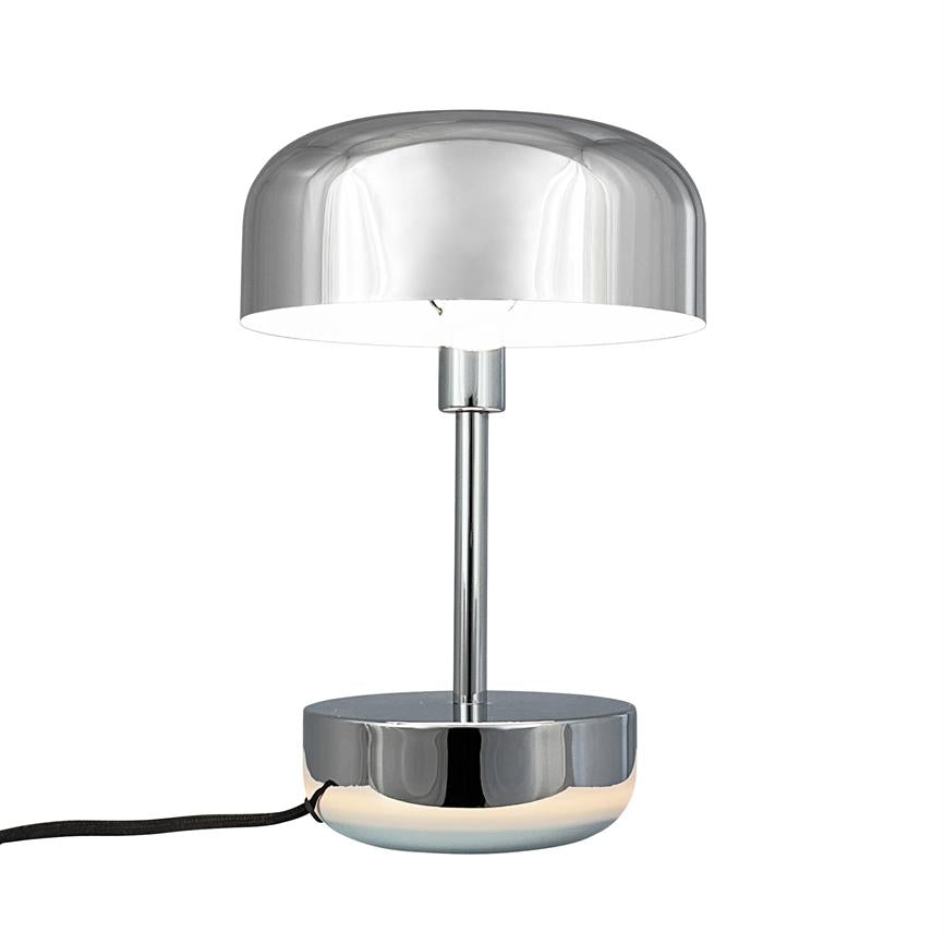 Bordlampe i blank krom står tændt, udsender blødt hvidt lys fra underside; slank stang og rund base med stofledning mod ren, hvid baggrund.