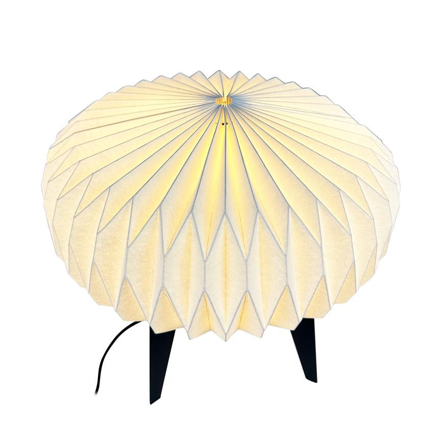 Runde, gefaltete Papierlampe mit gerillten, radialen Falten leuchtet sanft gelb, steht auf drei schwarzen Beinen mit sichtbarer Kordel auf neutralweißem Hintergrund.