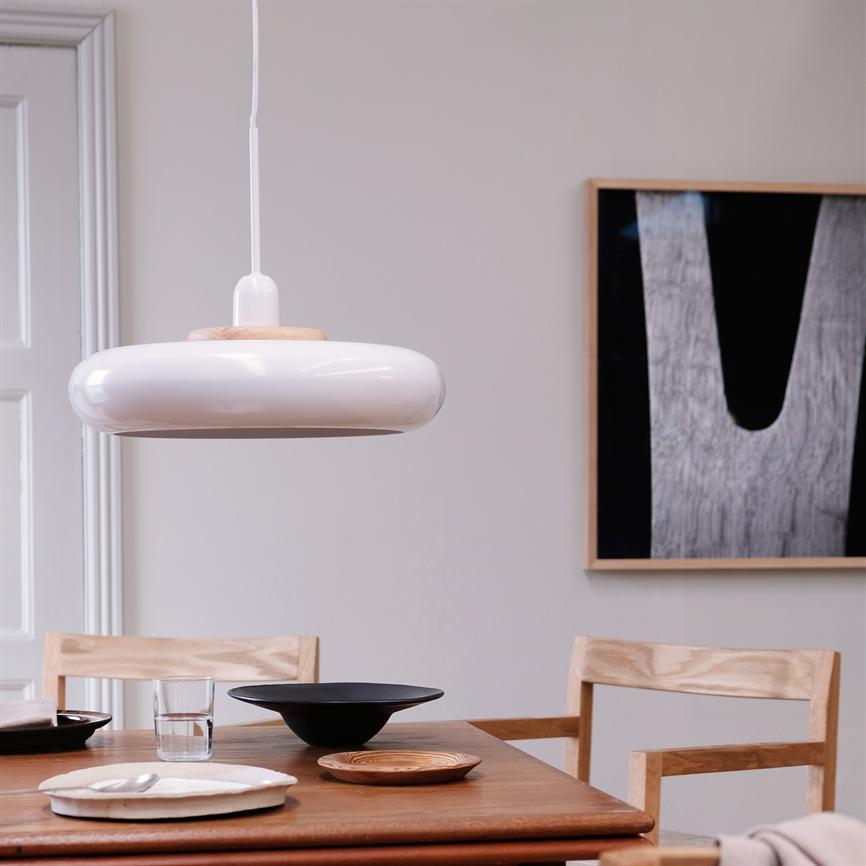 Runde weiße Pendelleuchte hängt über einem hölzernen Esstisch mit Schüsseln, Tellern und einem Glas; Zwei Holzstühle und ein abstraktes Schwarz-Weiß-Bild schmücken die Wand in einem hellen, minimalistischen Esszimmer.