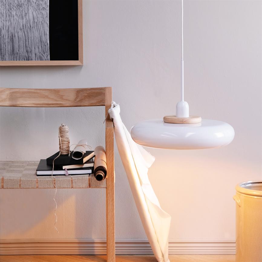 Eine weiße Pendelleuchte hängt und wirft sanftes Licht über einen Holzstuhl mit Büchern, Schnüren und Papierrollen; In einem hellen, minimalistischen Raum ist ein weißer Stoff nach unten drapiert.