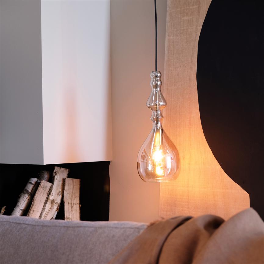 Eine dekorative Pendelleuchte aus Glas hängt und spendet warmes Licht. daneben in einer Nische gestapelte Birkenstämme, davor ein graues Sofa und abstrakte Wanddekoration.