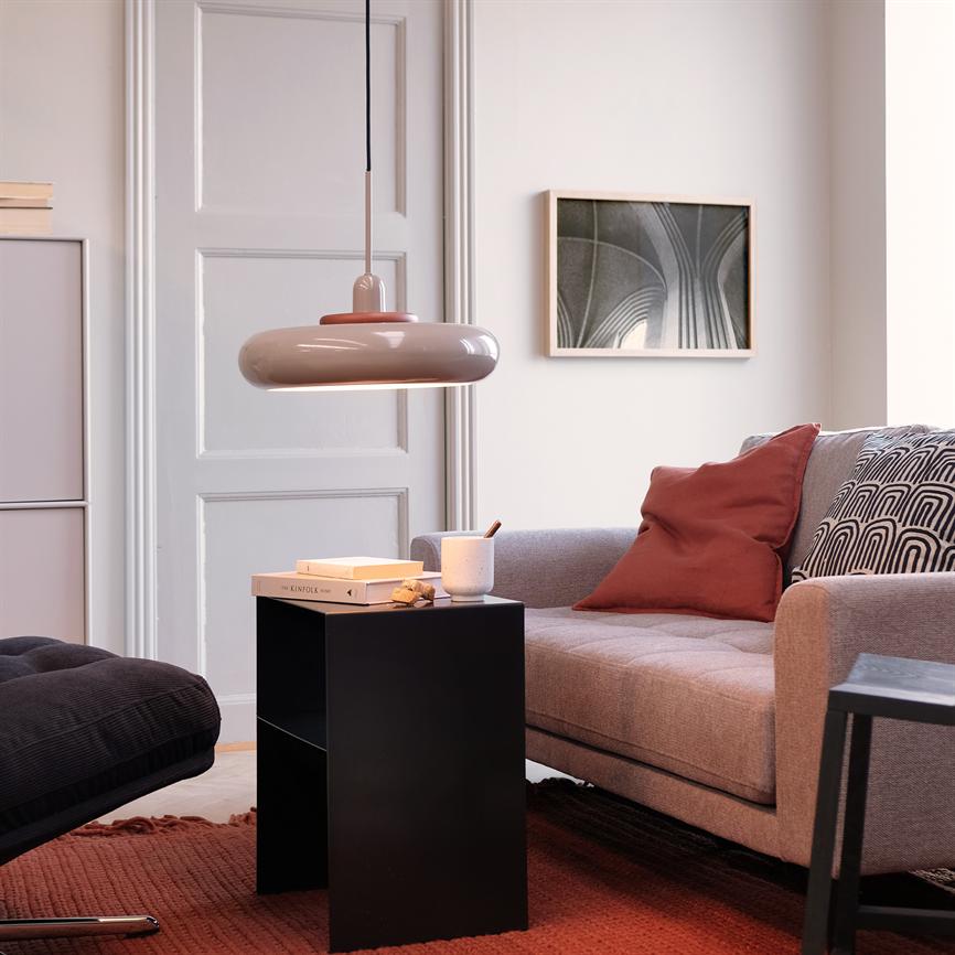 Kleiner schwarzer Beistelltisch steht mit einer Tasse und einem Buch mit der Aufschrift „KINFOLK“ vor einem grauen Sofa in einem hellen Wohnzimmer mit hängender runder Lampe, kastanienbraunem Teppich und Wandkunst.